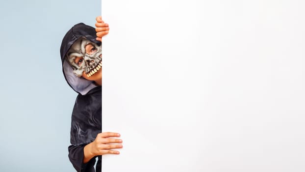 Happy Halloween. Cute little boy in a costume hiding behind white banner blank. Happy kid holding cardboard background with copy space.