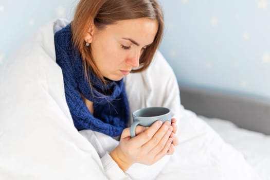 Sick woman covered with blanket holding cup of hot tea sitting on couch. Young woman is treated at home, took sick leave.