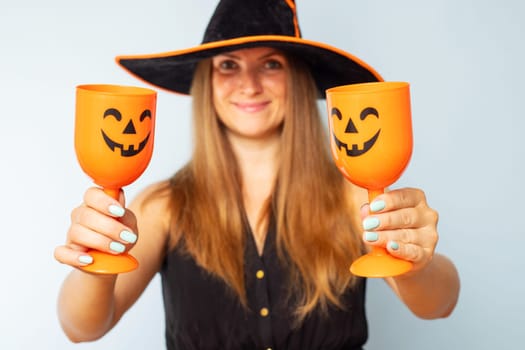 Happy Halloween. Happy brunette woman in halloween witch costume with black hat holding glasses of cocktails on a light background. Halloween party.