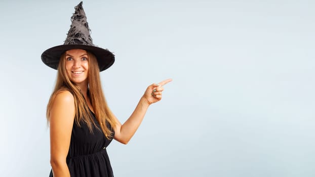 Woman in witch costume pointing with finger at copyspace side and looking at camera on blue background. Happy Halloween.