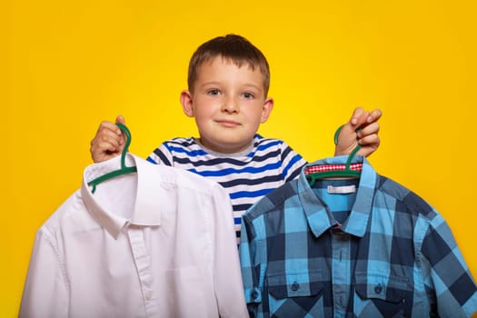 Cute little kid boy trying to decide between two shirt on yellow background. A boy holds two hangers with shirt in his hands.