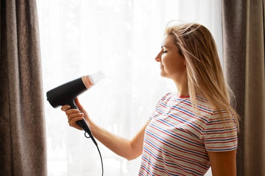 Beautiful young woman using hair dryer near the window at home.