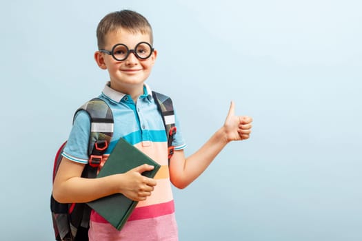 Back to school. Happy elementary school student in eyeglasses hugging book and showing a thumbs up gesture on blue background. Free space.
