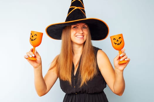Happy Halloween. Happy brunette woman in halloween witch costume with black hat holding glasses of cocktails on a light background. Halloween party.
