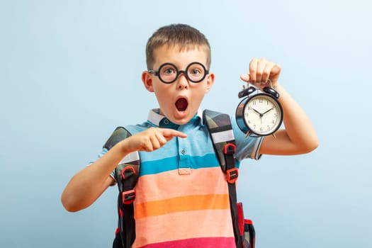 Shocked schoolboy in glasses with backpack holds an alarm clock in his hand and points at it with his finger, on blue background. Schedule and timing, back to school.