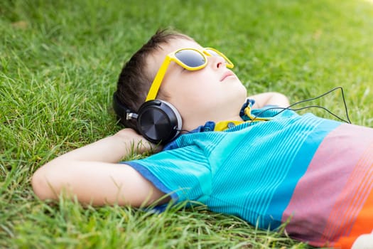 Happy boy in sunglasses lying on the grass in the park and listening to music in headphones on a sunny day.