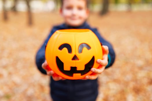 Child holds a bucket shaped like a halloween pumpkin jack o lantern in autumn park. Kid with jack-o-lantern. Halloween - traditional american holiday.