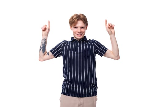 young European blond guy in a striped polo shirt with a tattoo shows his hand to the side on a white background.
