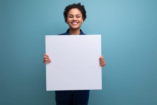 young successful woman with dark skin and black curly hair dressed in blue denim suit holding white blank poster for project showcase.