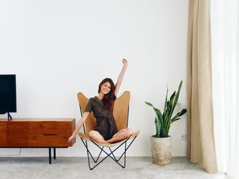 Woman sitting in a leather armchair near the window hands up smile , modern stylish interior Scandinavian lifestyle, copy space. High quality photo