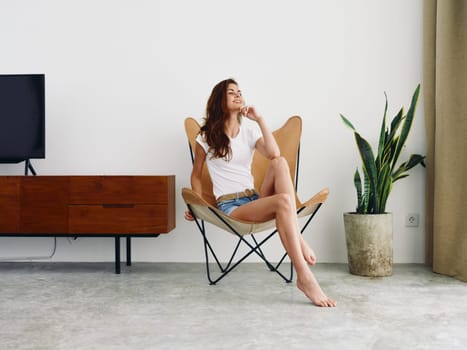 Woman sitting in leather armchair smile with teeth Lifestyle in white T-shirt and blue denim shorts, resting at home stylish modern interior with white walls, copy space. High quality photo