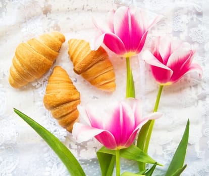 Three croissants and bright pink tulips on lace tablecloth.