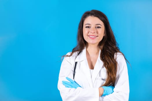 Long-haired woman crosses arms with smiling and delighted expression waiting for patient at blue wall. Young doctor with stethoscope enjoys working in health sphere