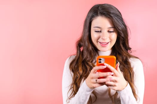 Brown-haired lady types messages to best-friends on smartphone. Young woman looks at correspondence with smiling expression in pink studio