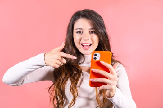 Young woman with delighted expression looks at smartphone pointing finger to wall. Brown-haired lady with amused expression at pink studio wall