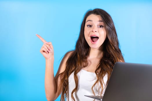 Young woman holding laptop advertises modern website at blue studio wall. Brown-haired lady with excited expression points to new boutique in shopping mall