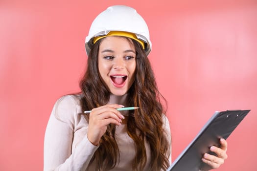 Woman wearing white hardhat writes information for building project on clipboard. Brown-haired architect with smiling and delighted expression enjoys job