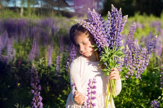 portrait of cute little happy two year old kid girl with bloom flowers lupines in field of purple flowers. nature outdoor.