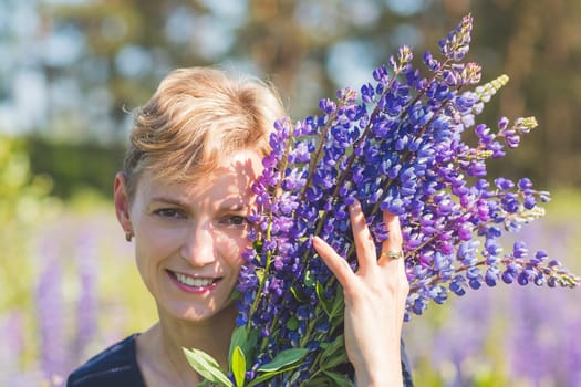 Portrait of young woman holding bouquet of lupin flowers walking in summer meadow.