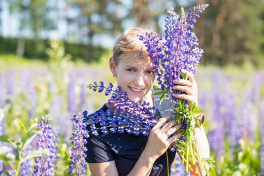 Portrait of young woman holding bouquet of lupin flowers walking in summer meadow.