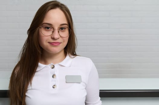 Confident close-up portrait of a young Caucasian woman wearing eyeglasses, a receptionist, administrator at reception desk in medical establishment. Hallway the emergency room or outpatient hospital