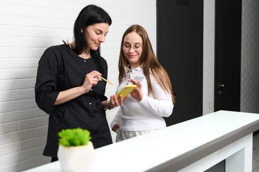 Medical staff. Young female doctor aesthetician with medical assistant using smartphone, checking list of appointments, standing behind a reception desk in modern white interior of a medical clinic