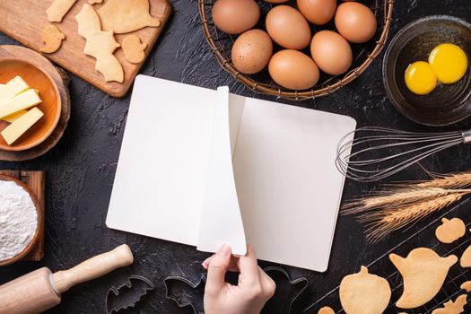 Woman is reading cookbook recipe of making Halloween cookies with baking ingredients, design concept of cooking class, top view, flat lay, overhead.