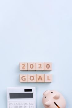 2020 goal, finance plan abstract design concept, wood blocks on blue table background with piggy bank and calculator, top view, flat lay, copy space.