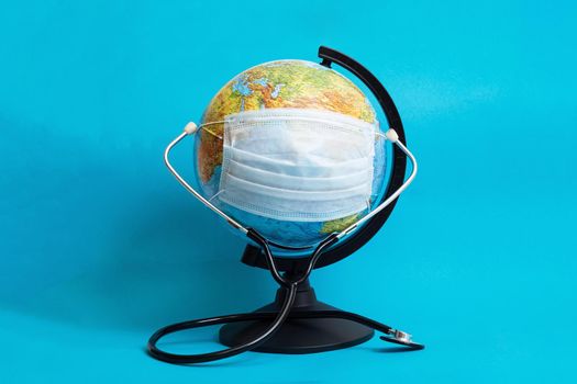 Globe in medical protective mask and stethoscope on blue background close up