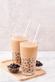 Tapioca pearl ball bubble milk tea, popular Taiwan drink, in drinking glass with straw on marble white table and wooden tray, close up, copy space.