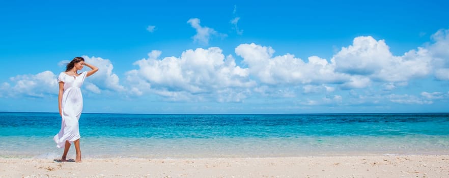 Woman in white dress walking on tropical beach, tropical sea on background