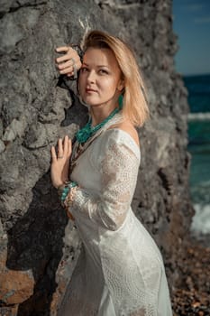 Woman white dress sea stones rocks.Middle-aged woman looks good with blond hair, boho style in a white long dress on beach jewelry around her neck and arms