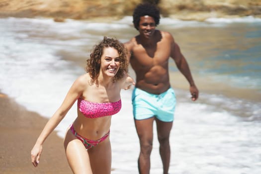 Cheerful multiethnic couple in colorful swimwear running on sandy shore while holding hands having fun together on resort