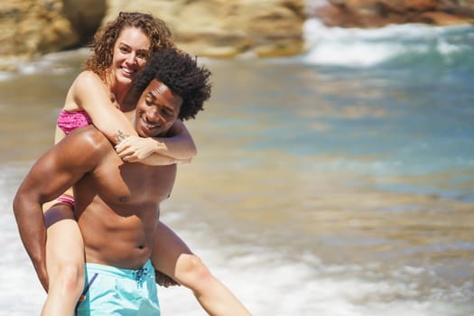 Cheerful young African American man with Afro hair giving piggyback ride to happy girlfriend on beach during honeymoon