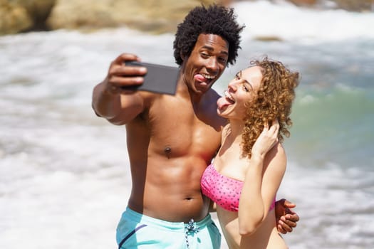 Cheerful multiracial couple in swimwear hugging and showing tongues while taking selfie on mobile phone for memory against waving ocean
