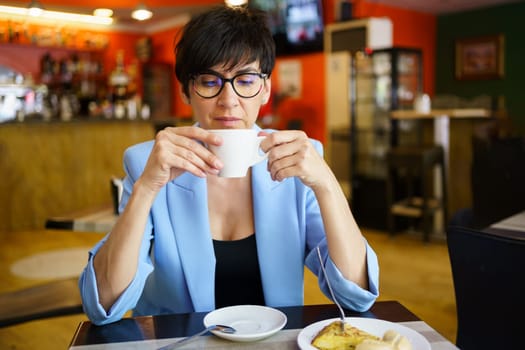 Focused mature female in eyeglasses and blue jacket drinking hot coffee from white ceramic cup while sitting at table in cafe
