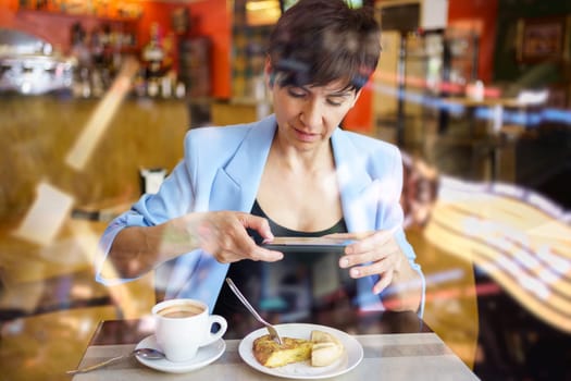Through glass view of young female in blue jacket, sitting at table with coffee cup and looking at screen of mobile phone while taking picture of snack in restaurant