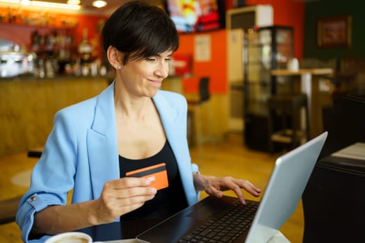 Focused middle age female entrepreneur in formal clothes sitting at table with laptop and making online payment with credit card while working in cafe