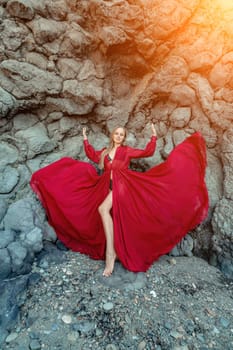 Red dress rocks woman. A blonde with flowing hair in a long flowing red dress stands near a rock of volcanic origin. Travel concept, photo session at sea.