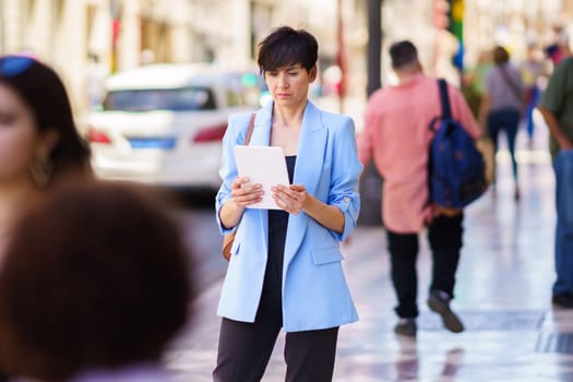 Serious woman in smart casual clothes with handbag standing on street and looking at screen of tablet while browsing in bright daylight