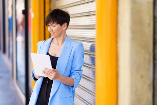 Positive female entrepreneur in formal clothes standing by silver rolling door with yellow column on street while browsing tablet in daylight