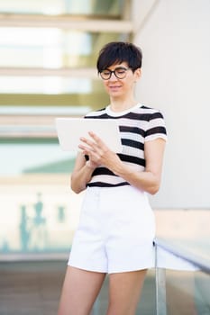 Positive female in casual outfit and eyeglasses standing in modern building and browsing tablet while leaning on railing