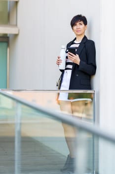 Pensive adult female in formal clothes with water bottle standing near glass wall of modern building and using mobile phone