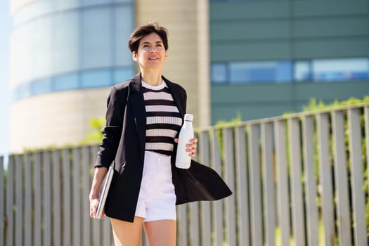 Positive female in trendy outfit with hand bottle to go smiling and holding laptop while walking along pavement near fence