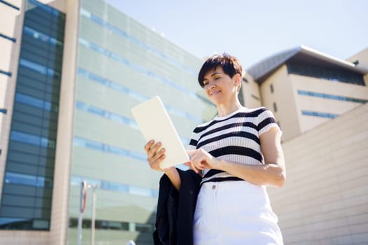 Low angle side view of female in casual clothes standing on street and reading notes in tablet while standing near modern building