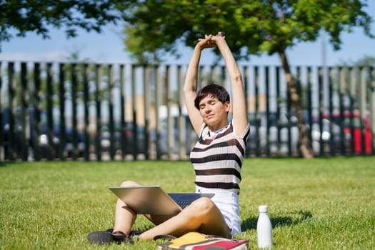Full body female freelancer in casual clothes sitting on grass with laptop and doing raised arms while relaxing during work remotely at park