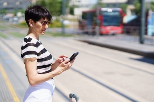 Focused adult female tourist in sunglasses and striped t shirt standing near suitcase and typing information on smartphone while waiting for transport on street