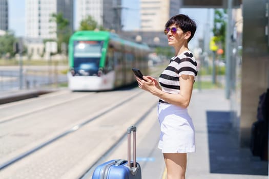 Side view of serious young female in trendy outfit and sunglasses standing near suitcase, and browsing mobile phone in train station with blurred background of train in city while looking away