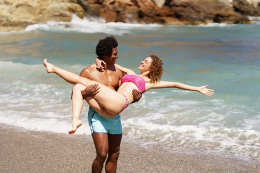Happy African American man raising woman with outstretched hands in swimwear while looking at each other during vacation against waving sea