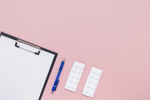 Flat lay of clipboard with paper, blue pen and blister pack of medicine pill on pink background for medical and healthcare concept